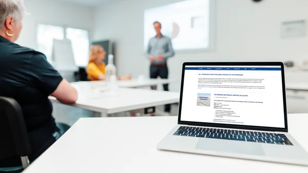 An open laptop can be seen in the foreground and a training room with three people can be recognised blurred in the background. Two people are sitting at tables and looking forwards to another person standing in front of a whiteboard.