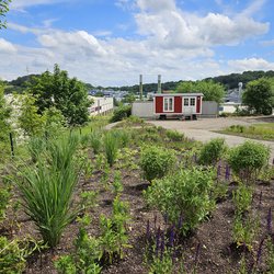 Auf dem Bild ist ein Garten mit einem angelegten Weg zu sehen. Im Hintergrund ist ein roter Bauwagen zu erkennen. Das Wetter ist sonnig mit blauem Himmel und ein paar weißen Wolken.