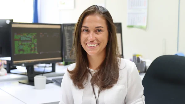 Frau in Büro lächelt, im Hintergrund sind Computerbildschirme.