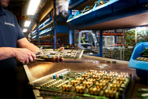 Person arbeitet an Elektronikplatine in Lagerhalle mit Regalen.