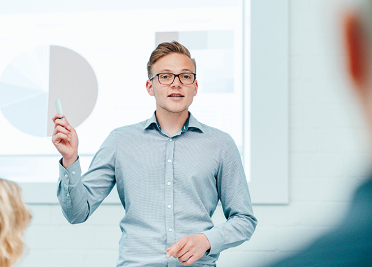 Auf dem Bild ist ein junger blonder Mann mit Brille in einem hellen Hemd zu sehen, der vor Publikum ewas erklärt.